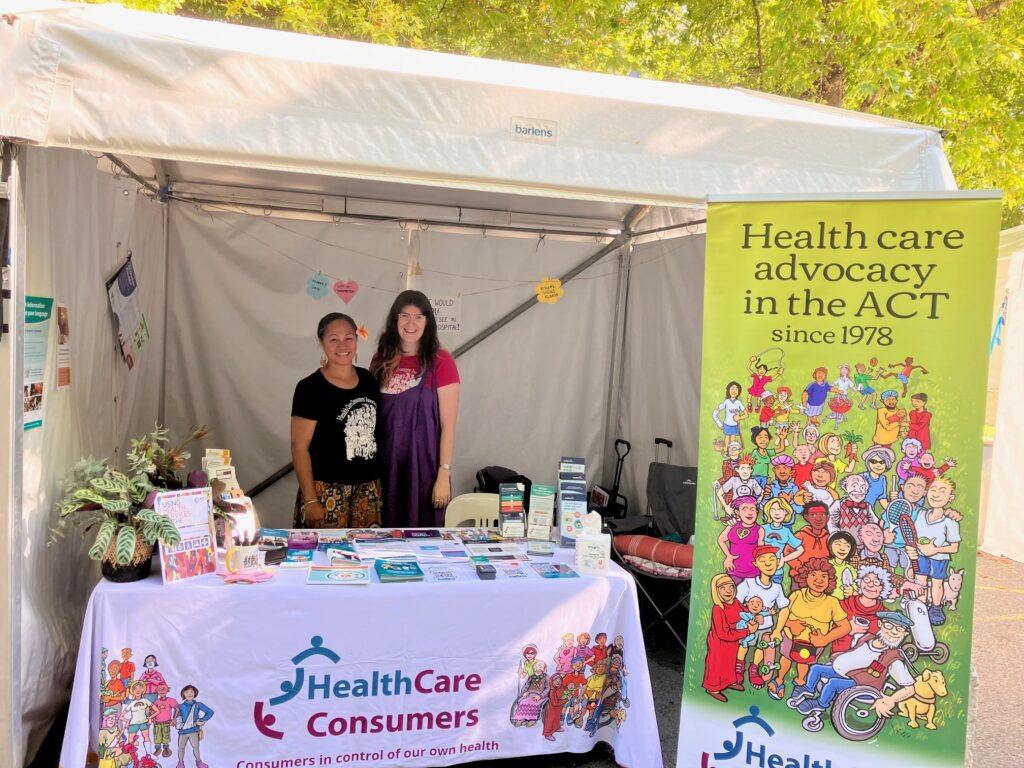 Our staff members Pene and Darcy stand smiling behind a table under a marquee. There are only a few paper decorations up at this point, but we have a bright banner and tablecloth.