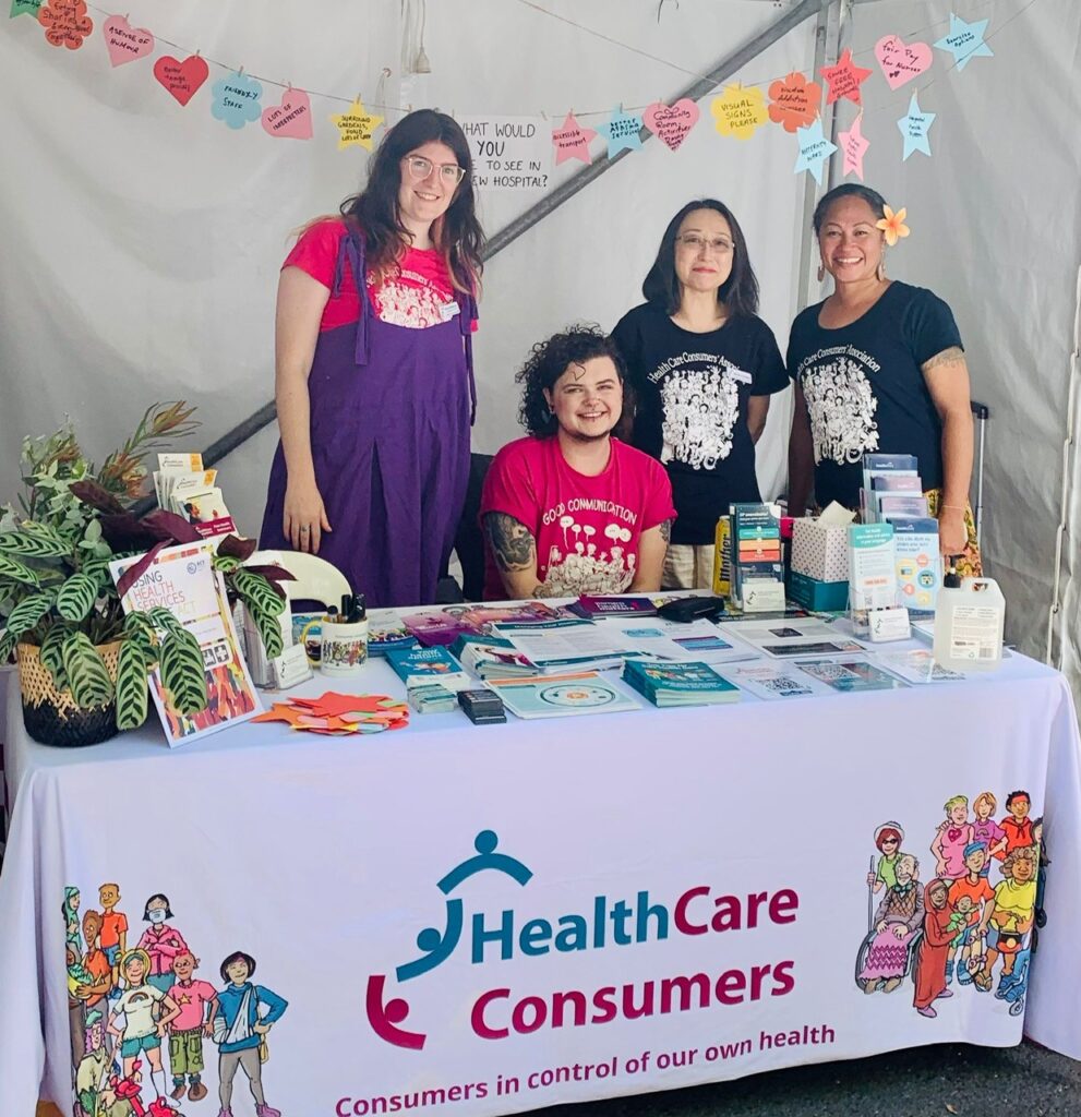 Staff members Darcy, C, Michiko, and Pene are behind a table at the stall. C is seated while the other three stand, and all are smiling at the camera.