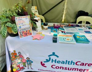 A photo of one corner of our stall, showing our colourful HCCA tablecloth, a stack of blank paper pieces to write on, some potted plants, and a range of paper resources with information for people to take with them.