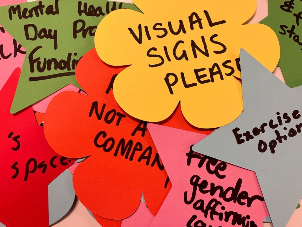 A pile of brightly colours paper shapes such as flowers and stars. Some of the things written on them with black marker include "visual signs please", "exercise options", "free gender affirming care", and something that ends with "... not a company."