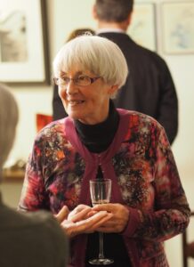 Kate Moore is pictured at some kind of event. She is a smiling woman with fair skin and short, loose, grey hair. She wears a colourful cardigan over a black turtleneck, is holding a champagne flute, and appears to be listening to someone just to the left of the photographer. The back and side of others at the event are visible, and there looks like framed art on a wall in the background.